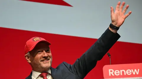 Getty Images A smiling Steve Reed in suit and tie waves, wearing a red baseball cap embroidered with the words "Build Baby Build!"