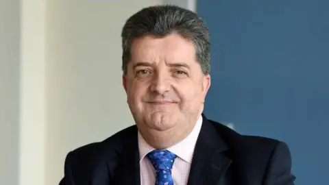 PA Media Ronnie Armour has dark hair swept back from his forehead and is wearing a dark suit jacket, white and pink checked shirt and blue tie with flouwers on it.