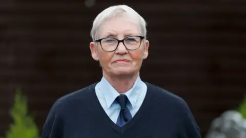 PA Media Elizabeth Hamilton has short grey hair and black-framed glasses and is wearing a navy v-neck jumper and tie