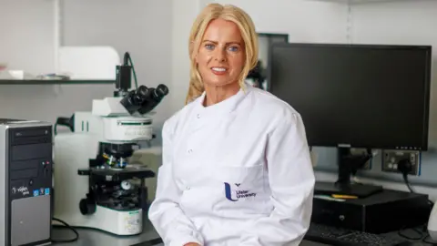 PA Media Tara Moore has long blond hair and is wearing a white Ulster University lab coat with a blue Ulster University logo on the left breast. To her right, behind her on a desk, is a large microscope while a desktop computer can be seen to her left.