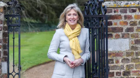 PA Media Hilary McGrady has long light blond hair and is wearing a grey coloured coat and yellow scarf while standing in a gateway - the gate is black wrought iron and on each side is a red brick wall. Behind her a green lawn can be seen. She is smiling.