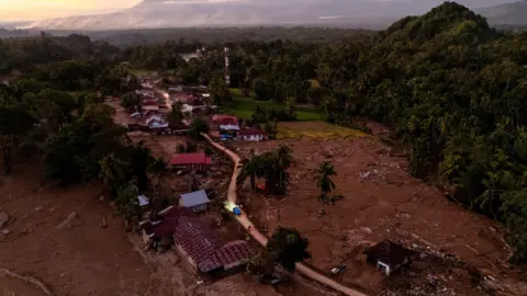 Reuters A drone view of vehicles driving through an area hit by deadly flash floods following heavy rains in Palembayan, Agam regency, West Sumatra province. The road cuts through fileds which appear to have been turned to mud, the houses are damaged. The area is surrounded by jungle, with a mountain in the background
