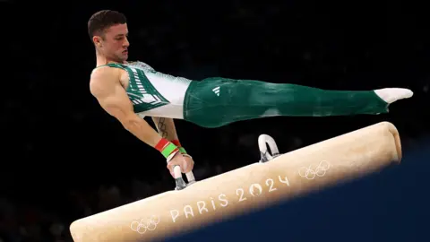 Reuters Rhys McClenaghan, who has short dark hair, competing in the 2024 Paris Olympics. He is wearing a green and white Ireland uniform and is swinging on the pommel horse